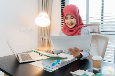 Ofiste dizüstü bilgisayarla çalışan tesettürlü Müslüman bir kadın, masadaki belgeler, planlama, analiz, mali rapor, iş planı, yatırım.