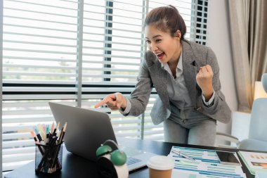 Mutlu Asyalı iş kadını zaferi ve olumlu ekonomik büyüme sonuçlarını kutluyor, dizüstü bilgisayarla belgeleri elinde tutuyor, ofiste çalışıyor.