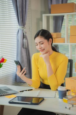 Ev ofisinde çalışan bir kadın dizüstü bilgisayar ve cep telefonu kullanıyor. Ürünler arka planda ambalaj için hazırlanıyor. KOBİ girişimcisi ya da serbest çalışan kavramı. Dikey resim.