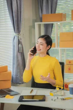 Ev ofisinde çalışan bir kadın dizüstü bilgisayar ve cep telefonu kullanıyor. Ürünler arka planda ambalaj için hazırlanıyor. KOBİ girişimcisi ya da serbest çalışan kavramı. Dikey resim.