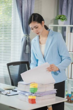 Masasında belgeler ve dosyalarla çalışan bir kadın var. Belgeleri düzenlemek, mali bilgileri gözden geçirmek. Masasında bir yığın belge tutarken önemli belgeleri inceliyor..