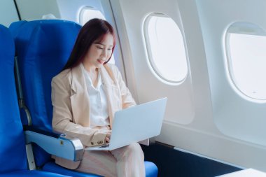 Genç bir kadın iş gezisi sırasında uçakta rahat bir şekilde seyahat ederken dizüstü bilgisayar kullanıyor. İş ve ulaşım kavramı.