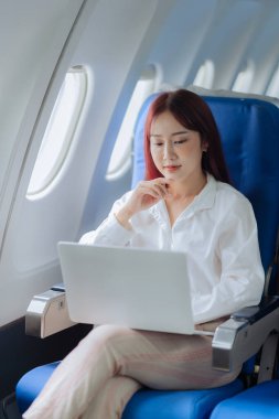 Genç bir kadın iş gezisi sırasında uçakta rahat bir şekilde seyahat ederken dizüstü bilgisayar kullanıyor. İş ve ulaşım kavramı.