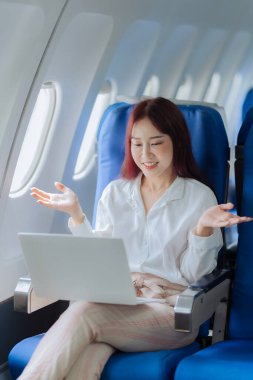 Genç bir kadın iş gezisi sırasında uçakta rahat bir şekilde seyahat ederken dizüstü bilgisayar kullanıyor. İş ve ulaşım kavramı.