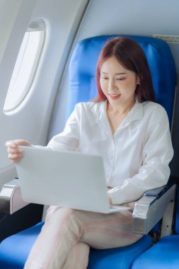 Genç bir kadın iş gezisi sırasında uçakta rahat bir şekilde seyahat ederken dizüstü bilgisayar kullanıyor. İş ve ulaşım kavramı.