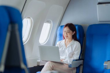 Genç bir kadın iş gezisi sırasında uçakta rahat bir şekilde seyahat ederken dizüstü bilgisayar kullanıyor. İş ve ulaşım kavramı.