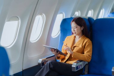 Asyalı iş kadını çalışmak için tablet kullanarak uçakla seyahat ediyor ve yeni başlatılan projenin kar ve finansmanı hakkındaki iş raporlarını kontrol etmek için kitap okuyor..