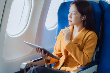 Asyalı iş kadını çalışmak için tablet kullanarak uçakla seyahat ediyor ve yeni başlatılan projenin kar ve finansmanı hakkındaki iş raporlarını kontrol etmek için kitap okuyor..