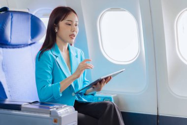 Asyalı iş kadını çalışmak için tablet kullanarak uçakla seyahat ediyor ve yeni başlatılan projenin kar ve finansmanı hakkındaki iş raporlarını kontrol etmek için kitap okuyor..