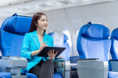 Asyalı iş kadını çalışmak için tablet kullanarak uçakla seyahat ediyor ve yeni başlatılan projenin kar ve finansmanı hakkındaki iş raporlarını kontrol etmek için kitap okuyor..