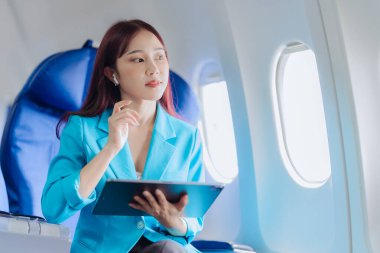 Asyalı iş kadını çalışmak için tablet kullanarak uçakla seyahat ediyor ve yeni başlatılan projenin kar ve finansmanı hakkındaki iş raporlarını kontrol etmek için kitap okuyor..