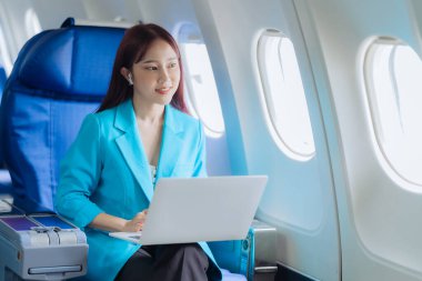 Genç bir kadın iş gezisi sırasında uçakta rahat bir şekilde seyahat ederken dizüstü bilgisayar kullanıyor. İş ve ulaşım kavramı.