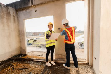 Asyalı endüstriyel erkek ve kadın mühendisler toplanıyor, makine planlarını analiz ediyor, inşaat alanındaki ofis masasında projeleri tartışıyorlar..