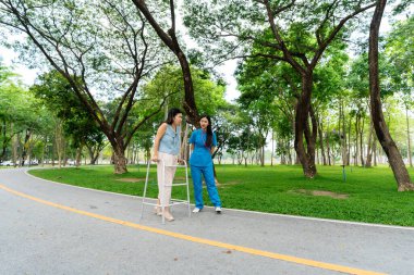 Asyalı hemşire bastonla yürüyen yaşlı kadına yardım ediyor. Parkta fizik tedavi gören yaşlı kadın hastalara yardım ve bakım yapıyor..