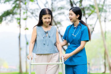 Asyalı hemşire bastonla yürüyen yaşlı kadına yardım ediyor. Parkta fizik tedavi gören yaşlı kadın hastalara yardım ve bakım yapıyor..