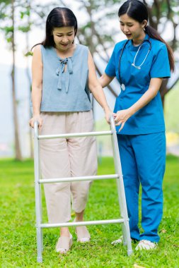 Asyalı hemşire bastonla yürüyen yaşlı kadına yardım ediyor. Parkta fizik tedavi gören yaşlı kadın hastalara yardım ve bakım yapıyor..