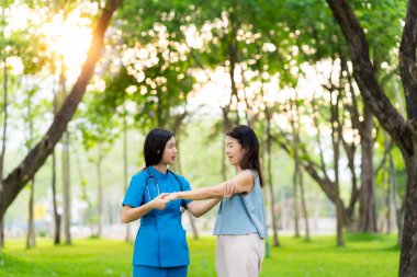 Asyalı kadın doktor parktaki yaşlı kadın hastayla ilgileniyor, hastaya parkta yürümesi ve dizlerini çalıştırması için yardım ediyor..