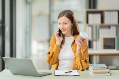 Girişimcilik ofisinde dizüstü bilgisayarla ve kitapla masada oturmuş cep telefonuyla konuşan gülümseyen genç kadın, pazarlama şirketi..