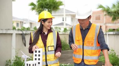 Yelekli ve miğferli iki Asyalı erkek ve kadın mühendis tablet üzerinde birlikte çalışıyor ve mimari yapı tasarımının planlarını tutuyorlar..