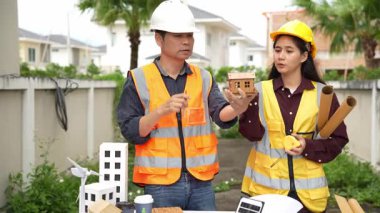 Yelekli ve miğferli iki Asyalı erkek ve kadın mühendis tablet üzerinde birlikte çalışıyor ve mimari yapı tasarımının planlarını tutuyorlar..