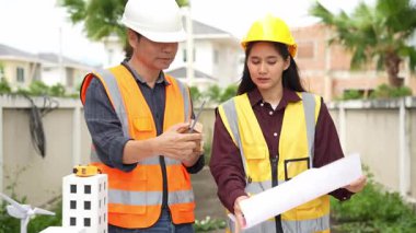 Yelekli ve miğferli iki Asyalı erkek ve kadın mühendis tablet üzerinde birlikte çalışıyor ve mimari yapı tasarımının planlarını tutuyorlar..
