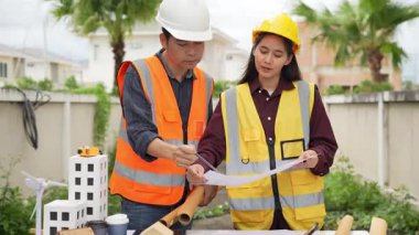 Yelekli ve miğferli iki Asyalı erkek ve kadın mühendis tablet üzerinde birlikte çalışıyor ve mimari yapı tasarımının planlarını tutuyorlar..