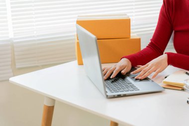 Önlüklü Asyalı kadın dizüstü bilgisayar ve fatura kutusu kullanıyor ve müşterilere paket hazırlamak için online siparişleri kontrol ediyor. Küçük işletme girişimcisine yakın çekim, girişim işi.