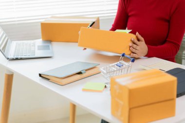 Önlüklü Asyalı kadın dizüstü bilgisayar ve fatura kutusu kullanıyor ve müşterilere paket hazırlamak için online siparişleri kontrol ediyor. Küçük işletme girişimcisine yakın çekim, girişim işi.