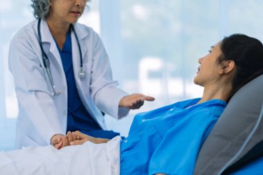 Elderly female doctor and Asian female patient are discussing something while sitting on hospital bed. Medical consultation concept.