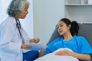 Elderly female doctor and Asian female patient are discussing something while sitting on hospital bed. Medical consultation concept.