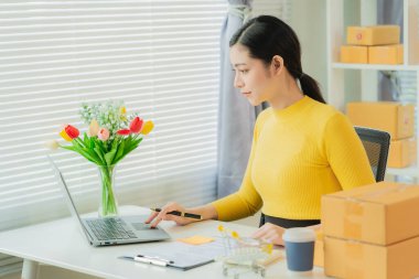 Cheerful Asian small business owner working in office with online sales and package delivery in home office.