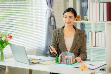 Real estate agent smiling while presenting a model home in the office holding a model home and working on a document about real estate concept.