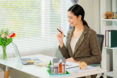 Real estate agent smiling while presenting a model home in the office holding a model home and working on a document about real estate concept.