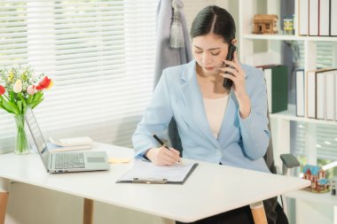 A white businesswoman with long hair in a formal dress is using a computer and talking on the phone, sitting in an office. A beautiful mind thinking freely.