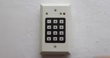 Bir kişi duvara monte edilmiş elektronik bir tuş takımına gizli bir kod giriyor. Video, kapının kilidini açmak için tuş takımı ile el etkileşimini gösteriyor.