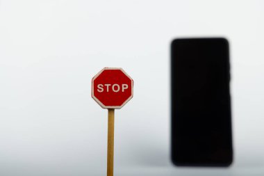 Kavramsal bir resim odak noktasında belirgin bir şekilde gösterilen bir oyuncak ahşap DUR işareti içerirken, bulanık bir akıllı telefon arka planda beyaz bir arkaplan karşısında görülebilir