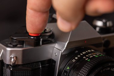 Koyu bir arkaplan ile zıtlaşan, panjur düğmesinde parmağı olan eski bir kameranın yakın çekim görüntüsü. Zamansız ve anımsatıcı.