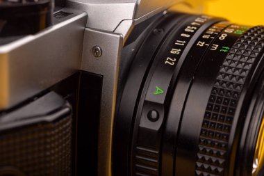 Eski bir kamera merceğinin ayrıntılı görüntüsü. Karmaşık tasarım ve işçiliğini vurguluyor. Parlak sarı bir zemin üzerine kurulmuş.