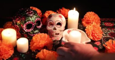Biri mumun içinde parlayan ve diğeri metalik çiçeklerle süslenen iki kafatası, kadife çiçekleri arasında yer alır. Video, mumları yerleştirip Da de los Muertos 'u onurlandırmanın sembolik eylemini yakalıyor..