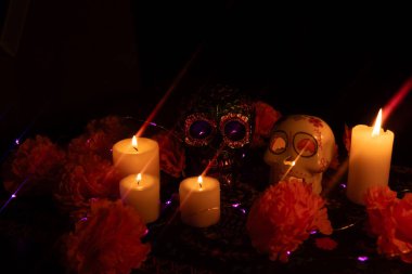 Close-up of two Day of the Dead skulls on a black backdrop: one decorated with metallic flowers and one with a candle burning inside. Marigold petals and lit candles surround the scene, with violet lights adding ambiance. clipart