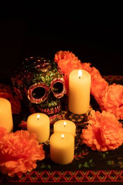 Metalik çiçeklerle süslenmiş, parlak turuncu kadife çiçekleriyle çevrili bir kafatasının dikey görüntüsü. Etrafında mumlar yanıyor, hepsi siyah bir arkaplan ve Ölü Masa Örtüsü Günü 'ne karşı..