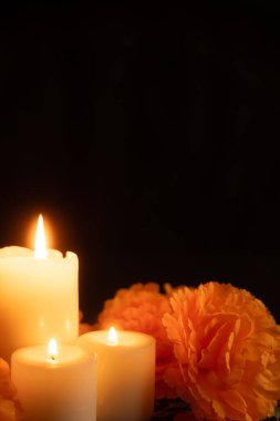 A vertical image with three candles glowing on the left, surrounded by orange marigold flowers. The scene is set against a black background with a Day of the Dead cloth, focusing on the candlelight.