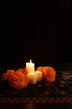 A vertical shot featuring three lit candles on the left, surrounded by vibrant orange marigolds. The scene is set against a black background with a Day of the Dead-themed tablecloth, illuminated only by candlelight.