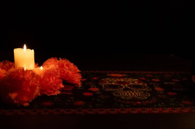 Parlak turuncu kadife çiçekleriyle çevrili, üç mumdan oluşan atmosferik bir çekim. Siyah arkaplan, Dia de los Muertos kumaşıyla geleneksel elementler ekleyerek mum ışığını vurgular..