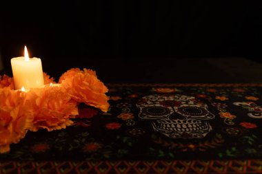 Solda, turuncu kadife çiçekleriyle çevrili, siyah bir zemin üzerinde titreşen üç mumun görüntüsü. Dia de los Muertos masa örtüsü mum ışığında havaya şenlikli bir dokunuş katar..