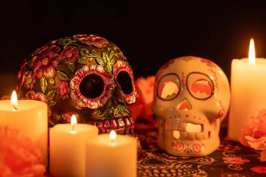 Karşıt özelliklere sahip iki kafatası: biri metalik çiçek süslemeleri ve biri mum ile. Etrafları kadife çiçekleri ve mumlarla çevrili. Hepsi de Dia de los Muertos desenli bir kumaş üzerinde..