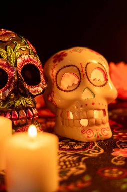 Bir Ölüm Günü masa örtüsünde iki kafatası bulunan dikey bir görüntü. Bir kafatası metalik çiçeklerle süslenirken, diğeri, içinde yanan bir mum bulunan içi kadife çiçekleri ve yanan mumlarla dolu, siyah bir arkaplan üzerine kurulmuş..