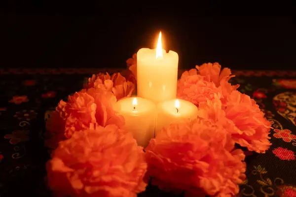 stock image A general view of three lit candles in the center, surrounded by vibrant orange marigolds. Set against a black background, the scene is illuminated solely by the candlelight, with a Day of the Dead tablecloth underneath.