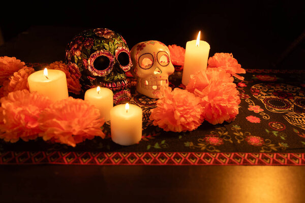 Вид сверху на череп, украшенный металлическими цветами, окруженный яркими оранжевыми ноготками и зажженными свечами. Черный фон и скатерть Day of the Dead завершают сцену.