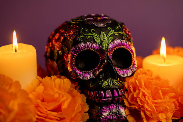 Крупный план замысловато детализированного черепа, украшенного металлическими цветами и окруженного ярко-оранжевыми ноготками. Две зажжённые свечи обрамляют сцену, отмечая Мексиканский день мертвых.